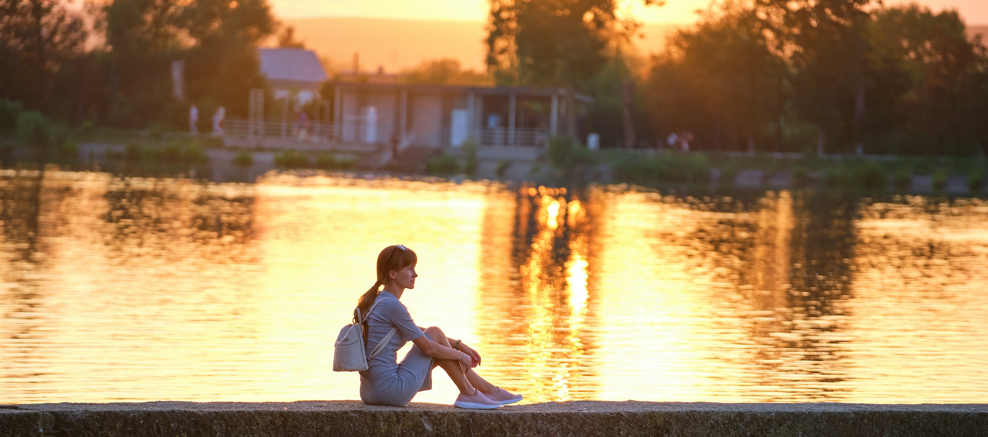 Side view of lonely woman sitting alone on lake shore on warm evening. Solitude and relaxing in